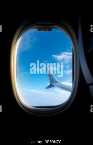 Blick aus dem Inneren eines Flugzeugs, der den Flugzeugflügel vor einem hellblauen Himmel mit Wolken zeigt, der während des Fluges in Reiseflughöhe erfasst wurde. Stockfoto