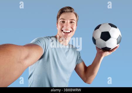 Lächelnder junger Mann mit Fußball nimmt Selfie auf seine Mobiltelefon über violettem Studiohintergrund Stockfoto