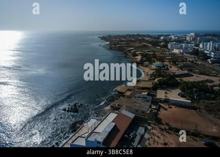 Ein Blick aus der Vogelperspektive auf Ngor, Dakar, zeigt eine Küste mit glitzernden Meereswellen, felsigen Ufern und einem kleinen Gebäude mit einem roten Dach. Die Szene umfasst Stockfoto