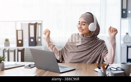 Freudige arabische Frau in Hijab, die Musik in Kopfhörern am Arbeitsplatz hört Stockfoto