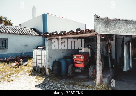 Rustikale Bauernhofsszene mit einem kleinen roten Vintage-Traktor, der unter einem verwitterten Schuppen geparkt wird, umgeben von traditionellen weiß getünchten Gebäuden, Kopfsteinpflaster-Gros Stockfoto