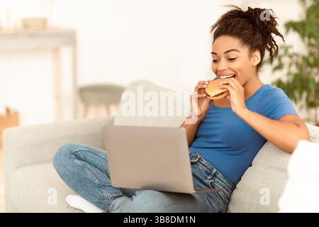 Schwarze Frau Genießen Ungesunde Burger Sitzen Auf Laptop Zu Hause Stockfoto