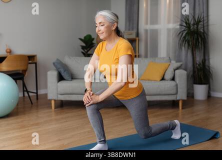 Positive sportliche Senior-Frau, die sich zu Hause fit hält, Ausfallschritte macht, im Wohnzimmer auf einer Yogamatte trainiert und lächelt Stockfoto