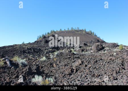 Die Landschaft zeigt ein krasses vulkanisches Gelände mit einem ausgeprägten Schlackenkegel im Hintergrund, umgeben von schwarzen Lavasteinen und verstreuten Sträuchern Stockfoto
