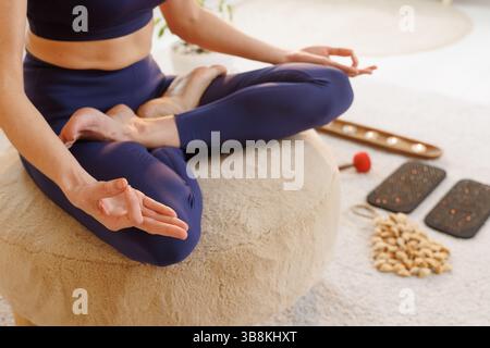 Nahaufnahme einer Frau in Lotus-Pose, die drinnen mit Mudra-Händen, Yoga-Werkzeugen und Heilungsgegenständen meditiert. Stockfoto