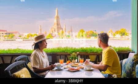 Ein Paar genießt ein friedliches Frühstück am Fluss in Bangkok, das lebhafte Gerichte und einen atemberaubenden Blick auf Wat Arun und üppiges Grün unter klarem Himmel bietet, was einen perfekten Morgen für einen Kurzurlaub sorgt. Stockfoto