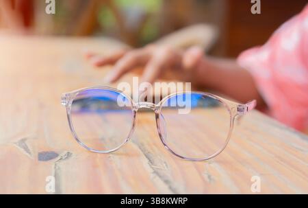 Nahaufnahme der Brille auf dem Holztisch mit verschwommenem Handbild mit Telefon dahinter. Nahaufnahme, selektiver Fokus. Stockfoto