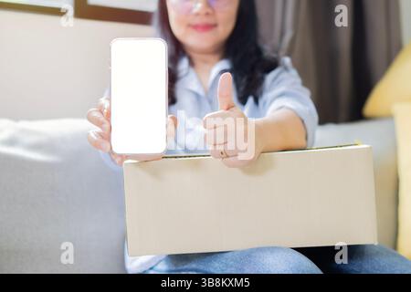 Asiatische Frau zeigt Daumen nach oben und benutzt Handy mit leerem Bildschirm Arbeitsplatz mit Karton. Auspacken des Pakets aus dem Versanddienst Stockfoto