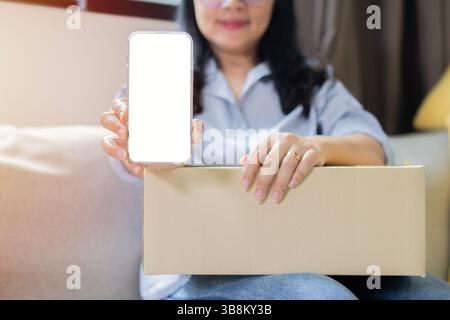Lächelnde schöne asiatische Frau, die Pappkarton hält und Hand zeigt Smartphone-Modell des leeren Bildschirms auf weißem Hintergrund Stockfoto