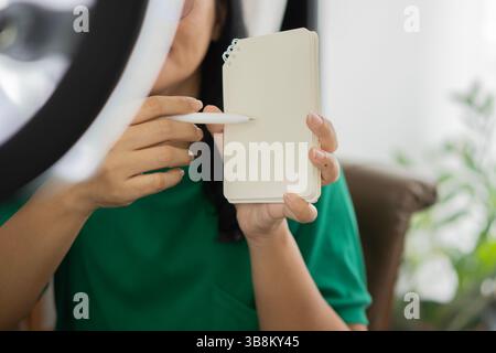 Junge asiatische Frau zeigt ihre Notiz und macht Foto- oder Videoinhalte für soziale Medien mit Smartphone und Licht einer Ringlampe. Stockfoto