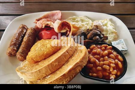 Komplettes englisches Frühstück auf einem Teller, mit Spiegeleiern, Speck, Würstchen, gebackenen Bohnen, Pilze, gegrillte Tomaten und Toast. Das Essen sieht herzhaft aus und Stockfoto