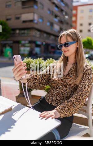Trendige Frau posiert mit Leopardenmotiv und Designer-Sonnenbrille und nimmt verspieltes Selfie am sonnigen Kaffeetisch im Freien auf Stockfoto