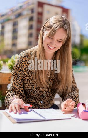 Junge blonde Frau, die studiert, Notizen am Bartisch macht, Hemd mit Leopardenmuster und fröhlichem Lächeln trägt Stockfoto
