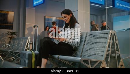 Überfülltes Flughafenterminal: Glückliche Frau mit Koffer, die nach der Sicherheitskontrolle in der Boarding Lounge sitzt, Flugticket und Dokumente überprüft, auf den Flug wartet. Passagiere, die an der Passkontrolle stehen. Stockfoto