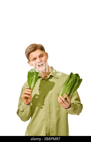 Fröhlicher Mann in hellgrünem Hemd, der knusprige Salatblätter mit lächelndem Gesicht und ausdrucksstarker Haltung vor weißem Studiohintergrund genießt Stockfoto