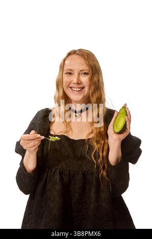 Lächelnde Frau in schwarzem Kleid, die Avocado und Löffel hält, genießt gesunden Biss mit freudiger Energie vor weißem Studiohintergrund. Stockfoto