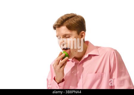 Junger Mann in rosa Hemd schließt die Augen und genießt säuerliche grüne Limette vor weißem Studiohintergrund. Stockfoto