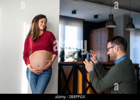 Lächelnde schwangere Frau zu Hause, während ihr Mann Selfie mit dem Handy macht. Romantischer Familienmoment eines jungen Paares, der Fotos macht. Ein glückliches Paar, das Stockfoto