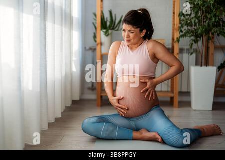 Junge schöne Frau im letzten Trimenon der Schwangerschaft, die Yoga-Hosen trug, unter unteren Rückenschmerzen litt und schmerzhafte Wehen hatte Stockfoto