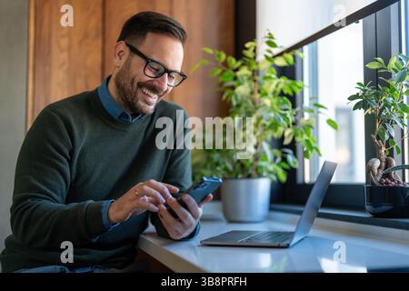 Selbstbewusster, lächelnder Geschäftsmann mit Smartphone im Heimbüro. Neue E-Mails werden geprüft. Videoanruf, Online-Kommunikationskonzept Stockfoto