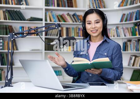 Eine Frau in einer Bibliothek, die ein Buch hält und Kopfhörer trägt, scheint Podcasting zu sein. Sie lächelt in die Kamera. Stockfoto