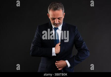 Ein reifer Geschäftsmann passt sein formelles Outfit an und konzentriert sich auf das Knöpfen seines Anzugs, während er in einem professionellen Studio nach unten blickt. Stockfoto