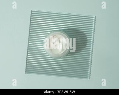 Kosmetische Creme oder Moustirizer auf transparenter gerippter Acrylplatte auf blauem Hintergrund. Offenes rundes Glasgefäß mit ästhetischer Wirbelcreme. Draufsicht FLA Stockfoto