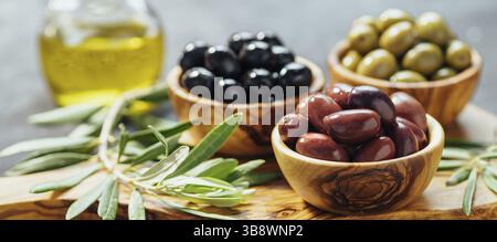 Set aus grünen, roten und schwarzen Oliven und Olivenöl auf grauem Hintergrund. Verschiedene Olivensorten in Olivenholzschalen und Olivenöl auf Holzschnitt b Stockfoto