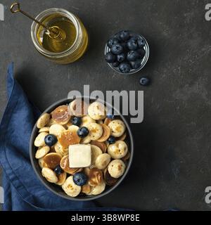 Trendiges Essen: Pfannkuchen, Müsli, Butter und Heidelbeeren. Haufen Mini-Cerealien-Pfannkuchen in Boul auf dunklem Hintergrund. Platz rechts für Text kopieren. Obere V Stockfoto