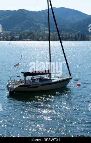 Schwarz-weißes Segelboot, das in glitzerndem Wasser mit orangener Boje und flatterndem Wimpel ankert, Gelassenheit am Berg unter blauem Himmel, nautische Ruhe. Stockfoto