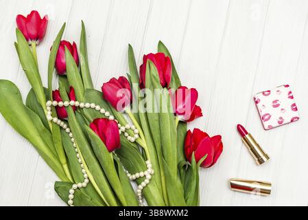 Blick von oben wunderschöner Blumenstrauß aus roten Tulpen in der Halskette, Lippenstift auf dem Hintergrund von weißem Holzbrett Stockfoto
