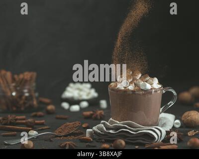 Von Hand bestreute Zimtpulver auf Glasbecher mit heißem Schokoladen-Kakaogetränk. Kopierbereich. Dunkler Hintergrund. Low-Taste. Winter Food and Drinks Konzept Stockfoto