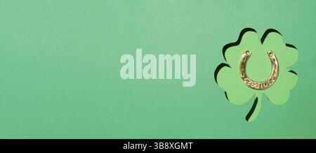 St. Patrick's Day Konzept. Grüner Papierhintergrund mit Schnittkontur aus Kleeblatt und goldenem Hufeisen in Blattform. Ansicht von oben oder flach liegend. Greetin Stockfoto