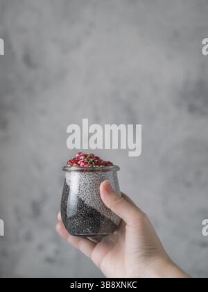 Chia-Pudding mit schwarzer Holzkohle und weißer Vanillemilch in der Frauenhand auf grauem Hintergrund. Weibliche Hand halten Glas mit Chia puding Serv Stockfoto