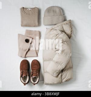 Damen warme leichte Daunenjacke, beiger Kaschmir-Pullover, rosa gestrickte Jogginghose, gestrickter Bini-Hut, braune Chukka-Wildlederstiefel aus Fell auf weißem Bettlaken. Stockfoto