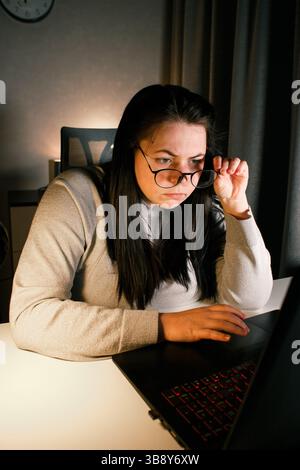 Junge Frau mit Brille, die spät in der Nacht am Laptop arbeitet, moderne Technologien nutzt und Online-Schulungen erhält. Fernlernkonzept. Stockfoto
