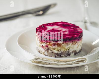 Geschichteter Salathering unter Pelzmantel auf dem Tisch. Portion traditionellen russischen Salats mit Hering, Rüben und anderem Gemüse auf weißer Leinentischdecke. Stockfoto