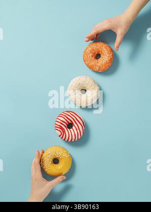 Zwei Hände greifen nach Donuts auf blauem Hintergrund, Draufsicht. Kreatives Layout mit köstlichen glasierten Donuts. Vertikale flache Lage mit Diagonale aus Donut Stockfoto