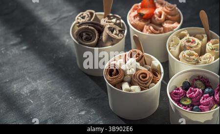 Gerolltes Eis in Kegelbecher auf dunklem Hintergrund. Verschiedene Eisrollen oder Thai Stil gerolltes Eis mit Kopierraum für Text oder Design. Banner. Natu Stockfoto
