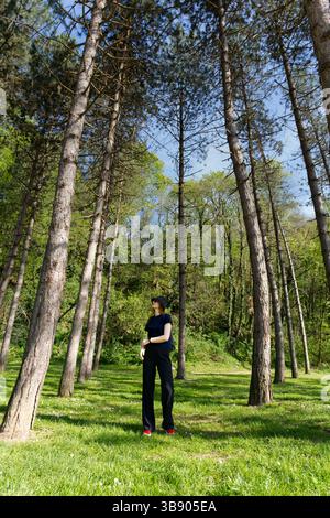 In Sonnenlicht getaucht, das durch hohe Kiefern filtert, blickt ein Mädchen in dunklem Outfit und Hut nach oben in einer ruhigen Waldlichtung mit einem grasbewachsenen Boden. Stockfoto