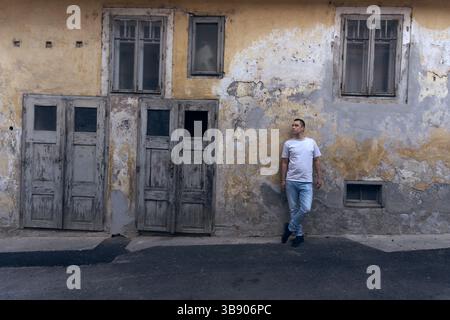 Das Bild zeigt ein altes, verwittertes Gebäude mit abblätternder Farbe und abgenutzten Holztüren. Ein lässig gekleideter Mann steht gegen die strukturierte Wand Stockfoto