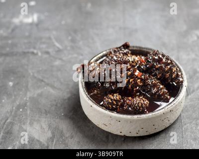 Köstliche Marmelade mit kleinen Tannenzapfen in einer kleinen Schüssel. Traditionelles sibirisches Dessert – Marmelade aus jungen Tannenzapfen auf grauem Hintergrund Stockfoto