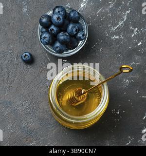 Gesunde Lebensmittel Zutaten - Heidelbeeren und Honig. Glasgefäß mit Honig und Dipper, frische Heidelbeeren in kleiner Schüssel über dunklem Hintergrund. Draufsicht oder f Stockfoto