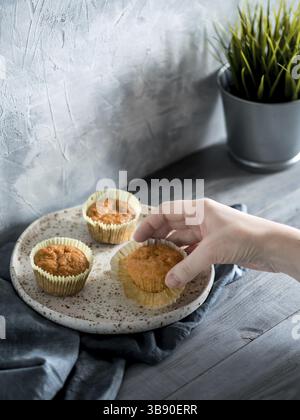 Hausgemachte Muffins auf Bastelplatte über grauem Holztisch. Karotten-Cupcake mit der Hand halten. Kopierbereich. Farblich abgestimmtes Bild im skandinavischen Stil Stockfoto