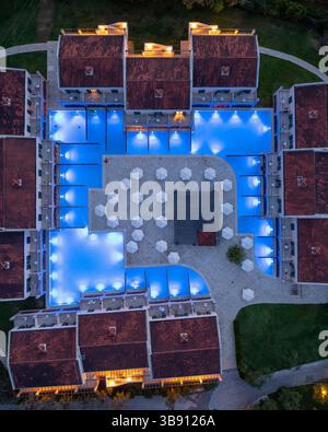 Blick aus der Vogelperspektive auf einen symmetrischen Hotelkomplex mit Pools, Sonnenschirmen und warm beleuchteten Gebäuden. Stockfoto