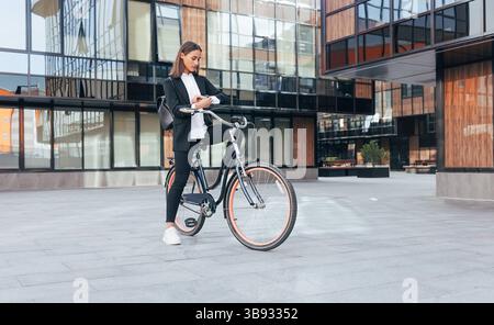 Junge Frau in Geschäftskleidung, die ihre Uhr überprüft, während sie mit einem Fahrrad im Innenhof eines Bürogebäudes steht. Frau in schwarzer Bürokleidung Dura Stockfoto