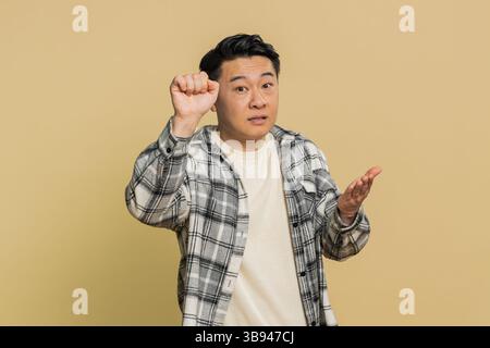 Klopfen-klopfen, wer da ist. Verwirrter aufgeregter asiatischer Mann, der an die Tür klopft und fragt, wer zu Hause ist, sich peinlich fühlt, keine Ahnung hat, ahnungslos und unsicher ist. Reifer Chinese auf beigefarbenem Hintergrund Stockfoto