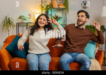 Fröhlich aufgeregtes junges indisches Paar tanzt High Five auf Sofa zu Hause sitzen. Lächelnde, glückliche, abwechslungsreiche Freundin und Freund, die ein Wochenende zusammen auf der Couch in der Wohnung genossen. Stockfoto