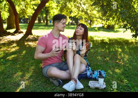 Ein glückliches junges Paar macht ein Picknick auf dem Gras in einem sonnigen Park und genießt gemeinsam Gebäck und Kaffee. Sie sehen sich an und lächeln Stockfoto