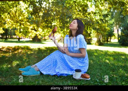 Eine glückliche junge Frau sitzt allein in einem sonnigen Park und genießt ein köstliches Picknick-Gebäck Stockfoto